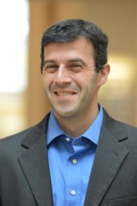 A man in a suit and blue shirt smiling.